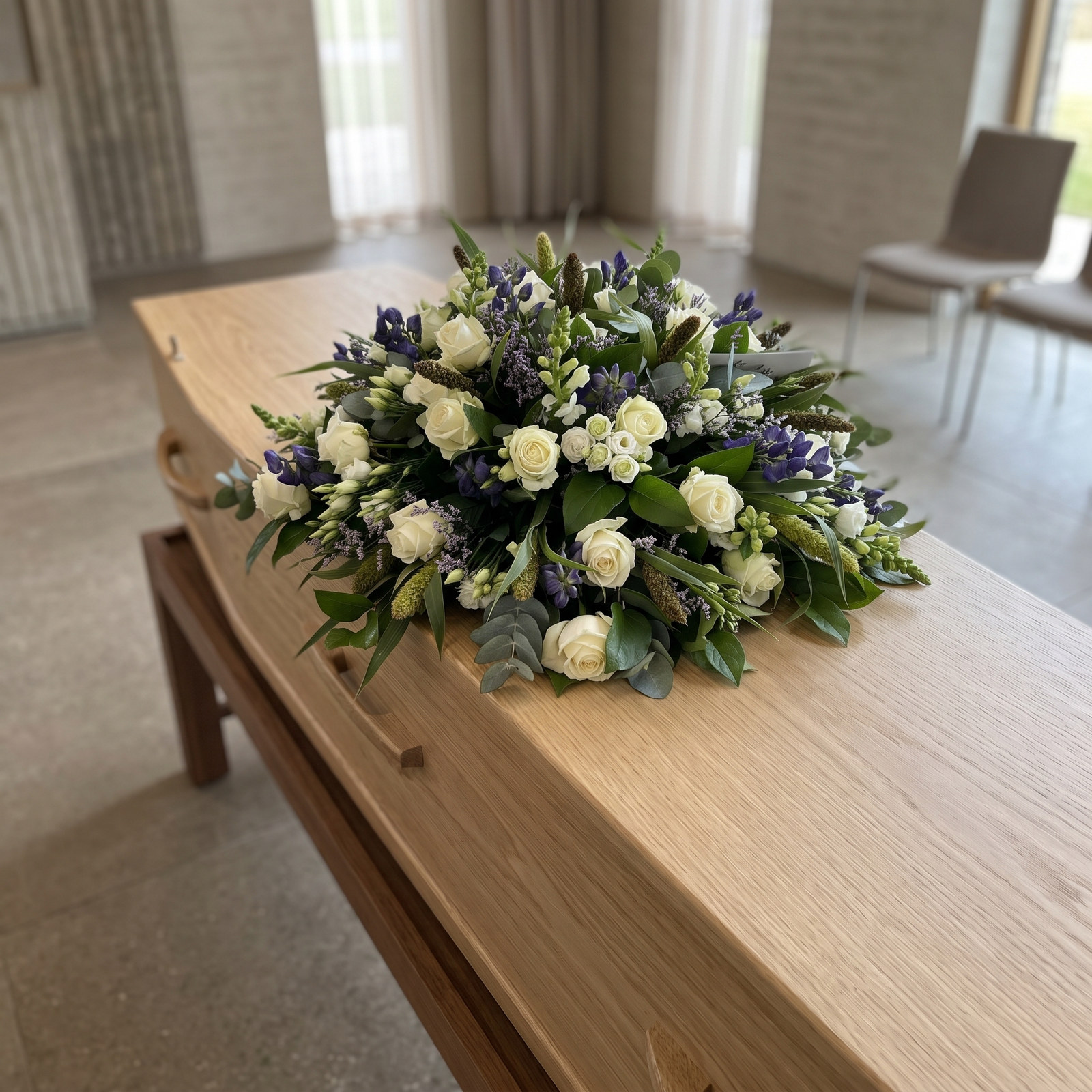 Rouwarrangement 'Afscheid nemen' met witte rozen met blauwe accenten en grijsgroen blad van bovenaf gezien op een lichte eiken kist