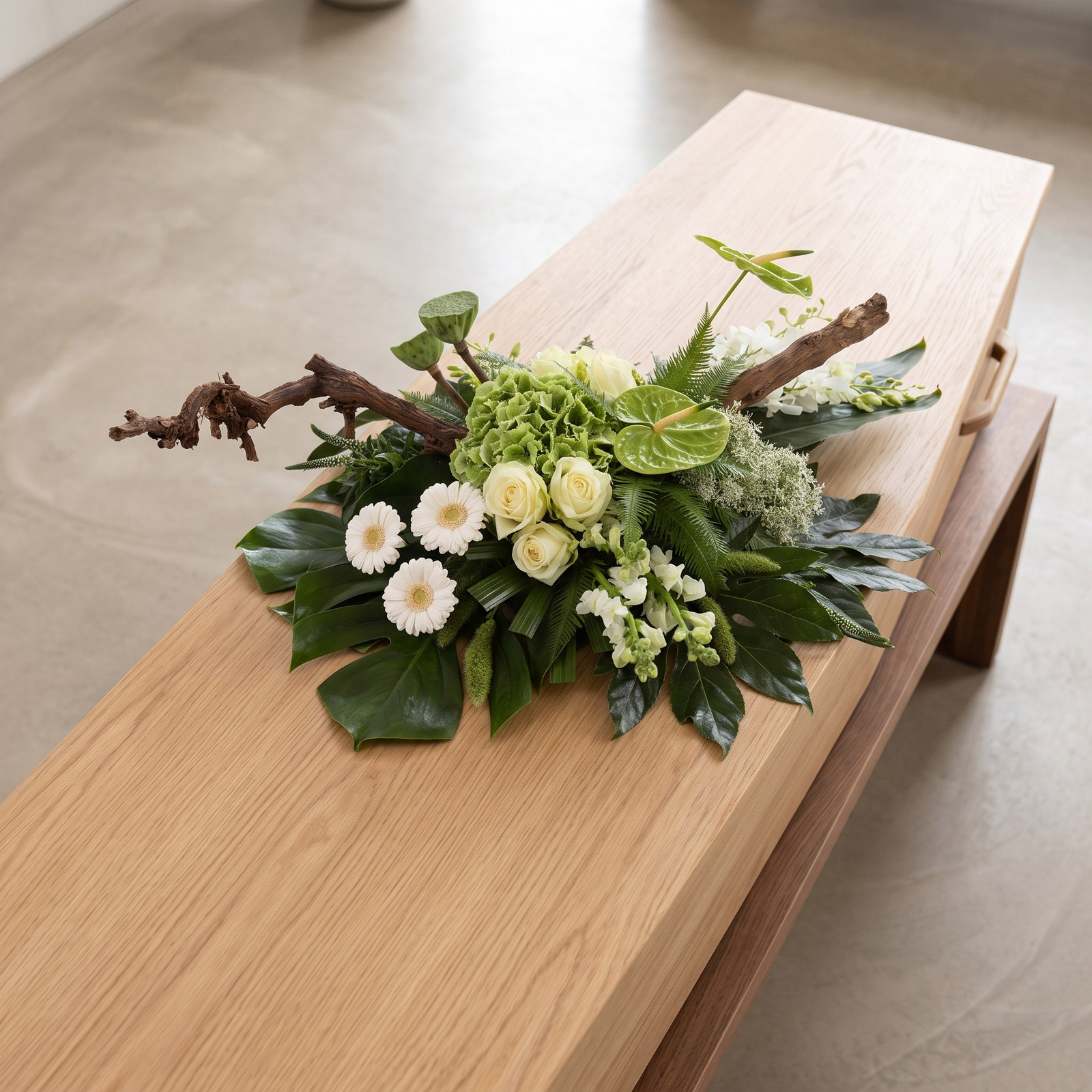 Rouwarrangement 'Ik mis je' met witte gerbera's en witte bloemen op grof blad met tak van bovenaf gezien op een lichte eiken kist